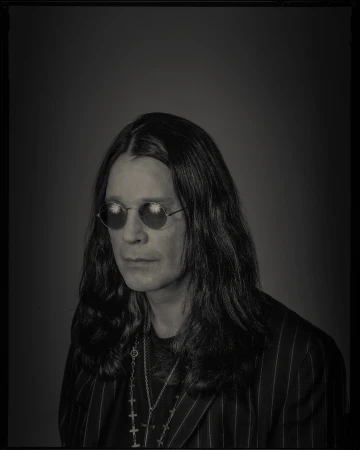 Black and white portrait of adult male celebrity, Ozzy Osbourne, wearing sunglasses, and a pinstripe blazer against a solid background.