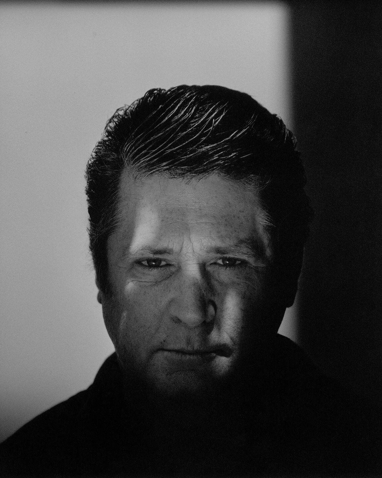 Monochrome portrait of a mature adult male with a serious expression, partially shadowed face, and slicked-back dark hair against a white background.