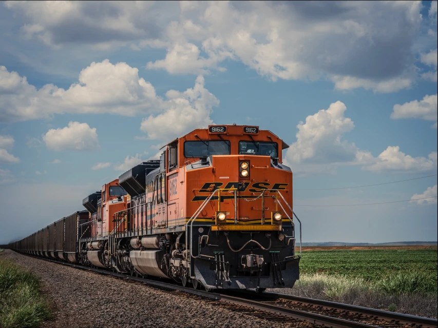 An orange BNSF freight train traveling on a track through a rural landscape with green fields under a blue, partly cloudy sky.