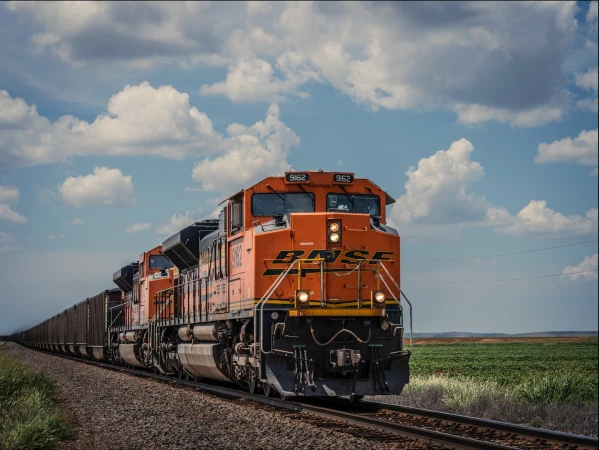 An orange BNSF freight train traveling on a track through a rural landscape with green fields under a blue, partly cloudy sky.
