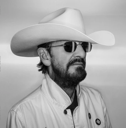Black and white portrait of Ringo Starr wearing a wide-brimmed cowboy hat, tinted sunglasses, and a collared jacket against a grey backdrop.