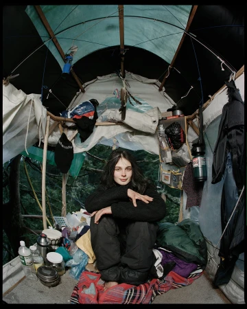 Young adult in a homemade indoor tent surrounded by gear, with a forest backdrop, creating an intimate, unique, alternative living space.