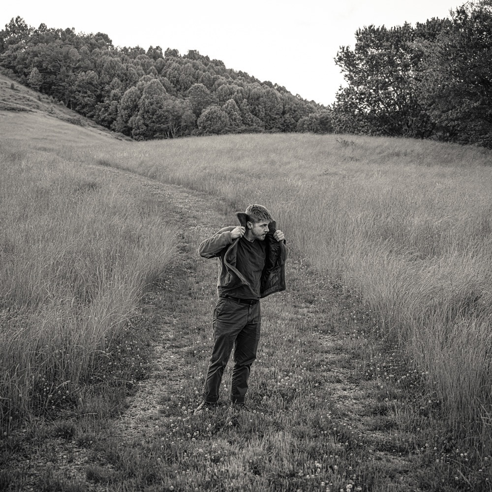 Monochromatic portrait of a man standing in a grassy field, putting on a jacket with a hill and trees in the background.