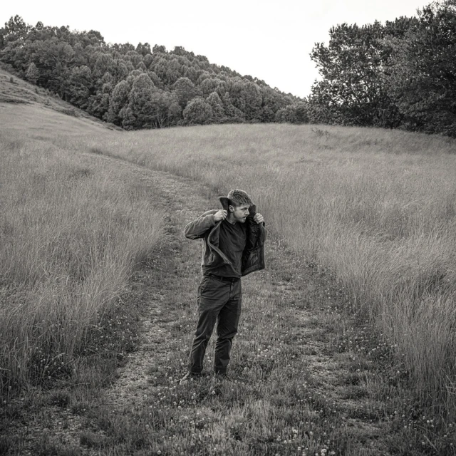 Monochromatic portrait of a man standing in a grassy field, putting on a jacket with a hill and trees in the background.