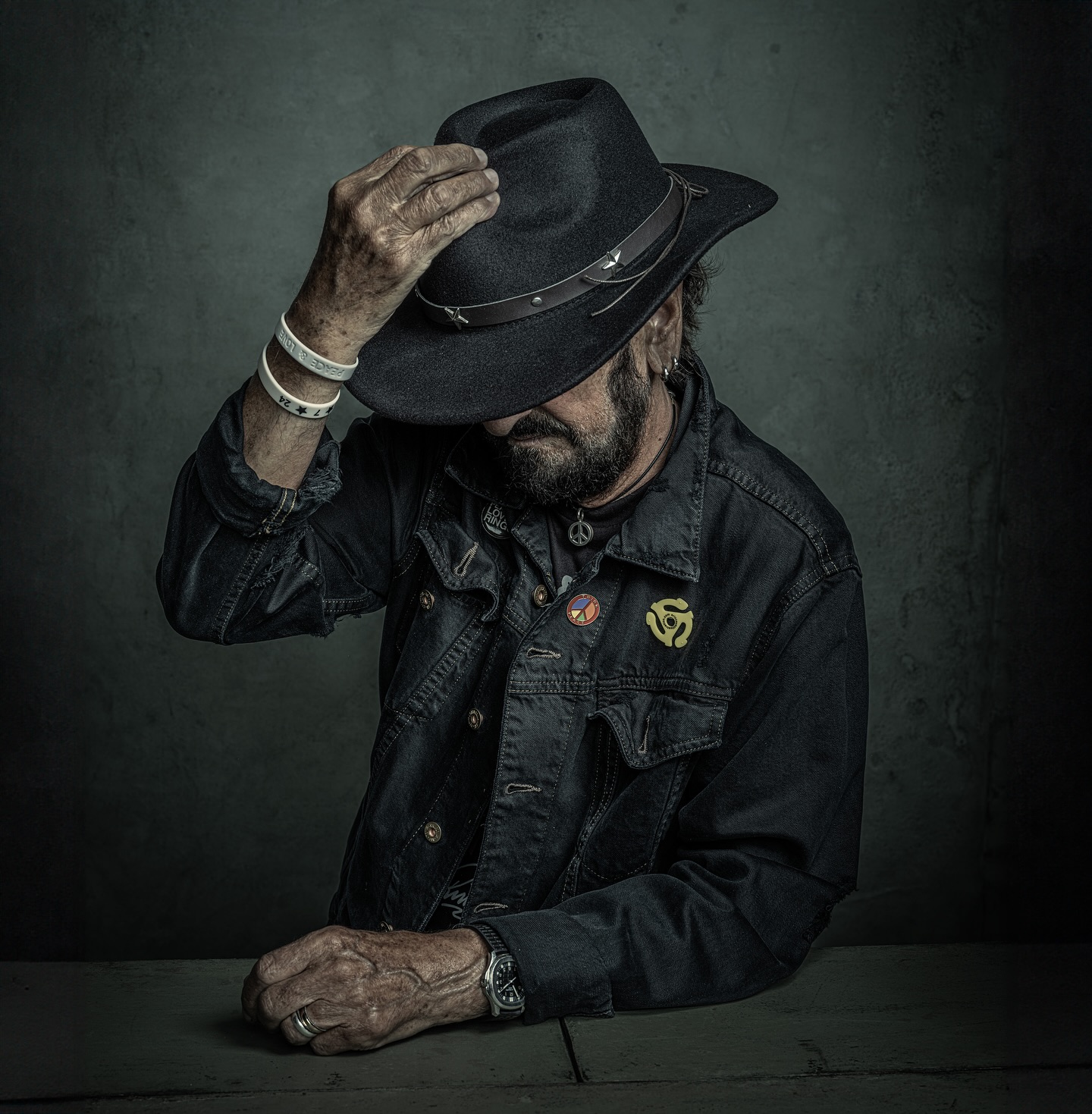 Portrait of a mature man wearing a black denim jacket and a hat, posing indoors with his hand near the brim of the hat.