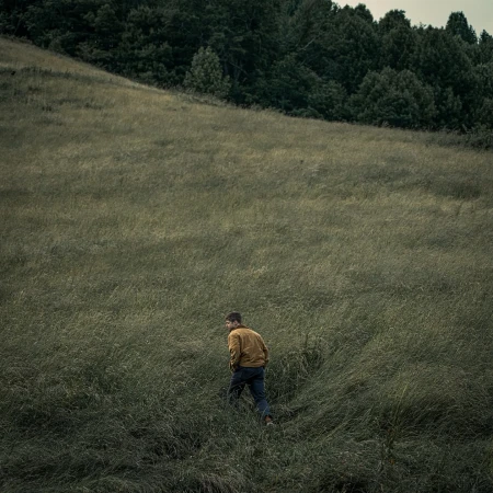 A man in a mustard jacket and dark trousers walks through a vast tall grass field toward a dense treeline on a hillside.