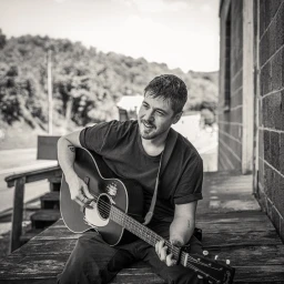 Monochrome portrait of a white man in his 30s sitting on a porch playing an acoustic guitar