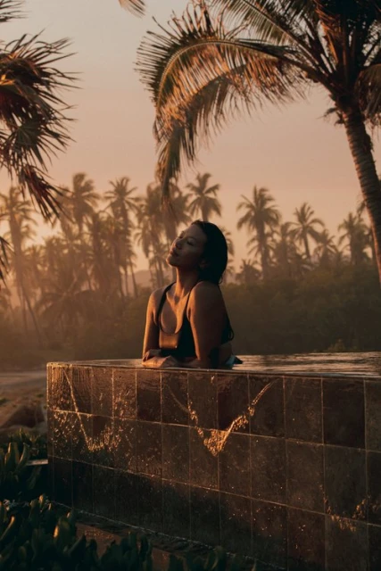 A woman relaxes in a pool surrounded by palm trees at sunrise. She is silhouetted against the warm, golden light.