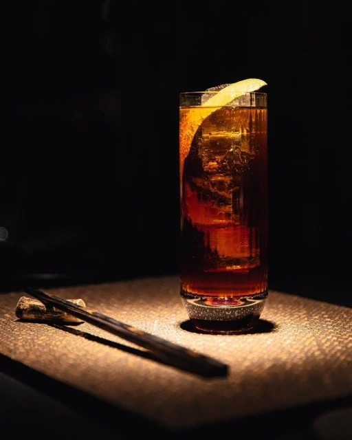 A highball glass of dark amber cocktail with ice, garnished with a lemon peel, set against a dark background, illuminated by focused lighting.