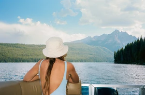 Woman in a light blue swimsuit and white hat sits on a boat, looking out at a lake surrounded by green trees and mountains under a blue sky.
