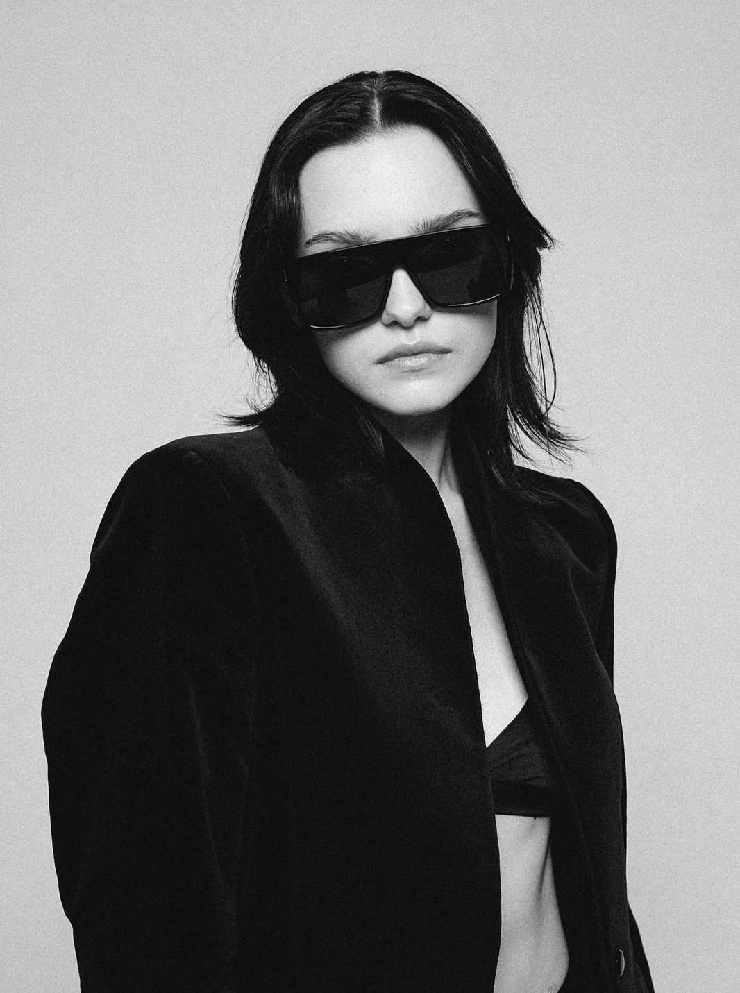 A woman with dark hair poses in a black jacket and sunglasses against a neutral background, creating a stylish portrait.