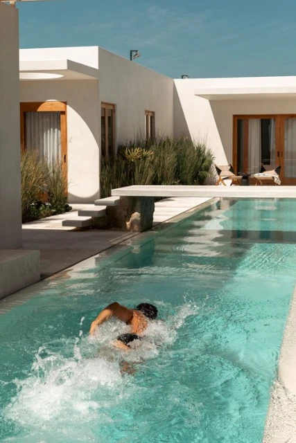Person swimming in an outdoor pool next to a modern white building with plants and patio furniture, sunny day, blue sky and water.