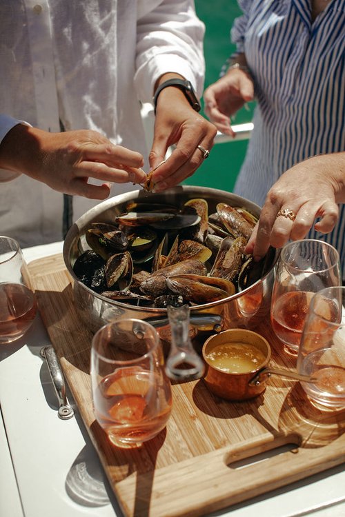 People sharing mussels from a silver pot on a wooden board, served with wine glasses on a bright sunny day outdoors.