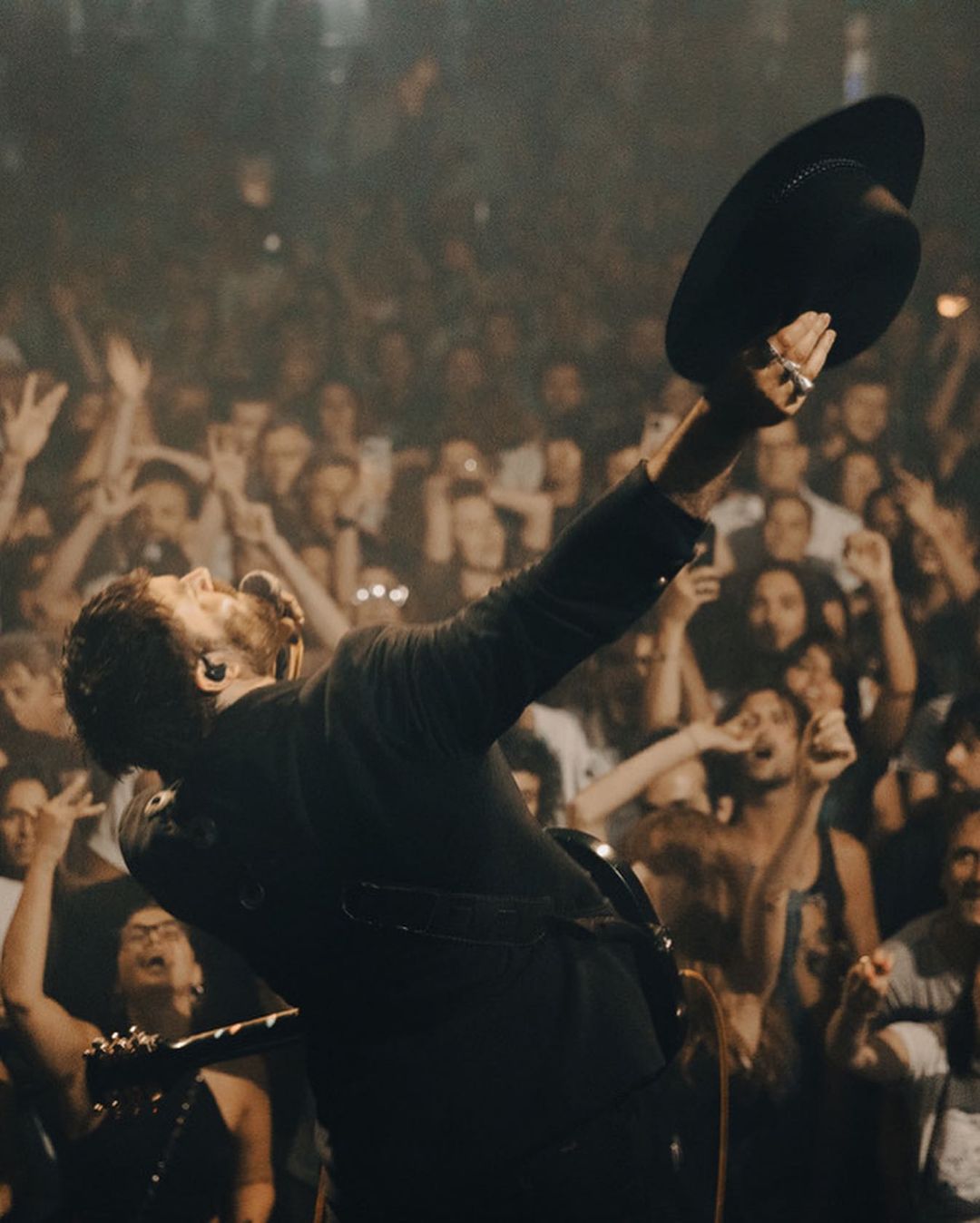 A singer in a black suit raises a hat on stage in front of a crowd, illuminated by concert lighting, creating a dynamic scene.