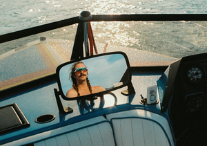 Man with braided long hair and mustache reflected in a boat mirror on sparkling water