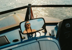Man with braided long hair and mustache reflected in a boat mirror on sparkling water