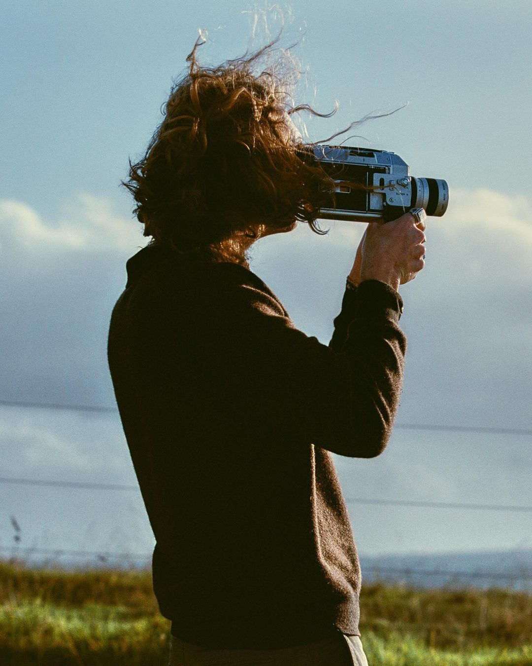 Person with long hair filming with vintage camera against sky and grassy field, wearing brown sweater outdoors in natural lighting.