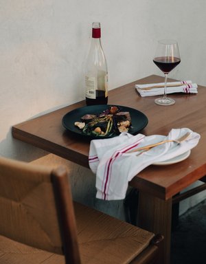 Still life food photography: a dark plate of steak and vegetables on a wooden table, wine glass and bottle, lit by natural light.