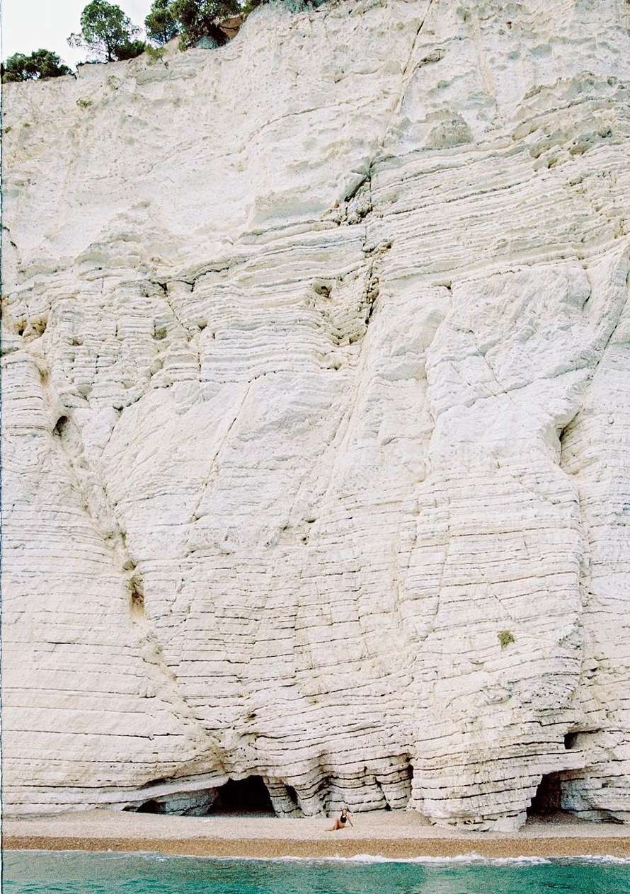 Striking view of a vast white rock cliff face with layered geological formations, small trees atop, and a petite turquoise sea below.
