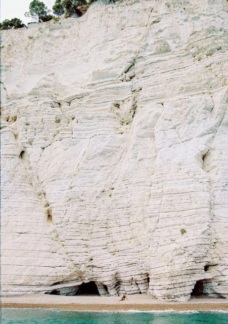 Striking view of a vast white rock cliff face with layered geological formations, small trees atop, and a petite turquoise sea below.