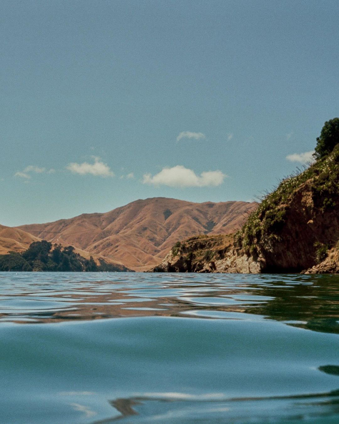 Water-level shot of calm blue water with gentle ripples, showing distant brown mountain ranges and a rocky, vegetated coastline.