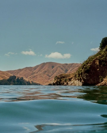 Water-level shot of calm blue water with gentle ripples, showing distant brown mountain ranges and a rocky, vegetated coastline.