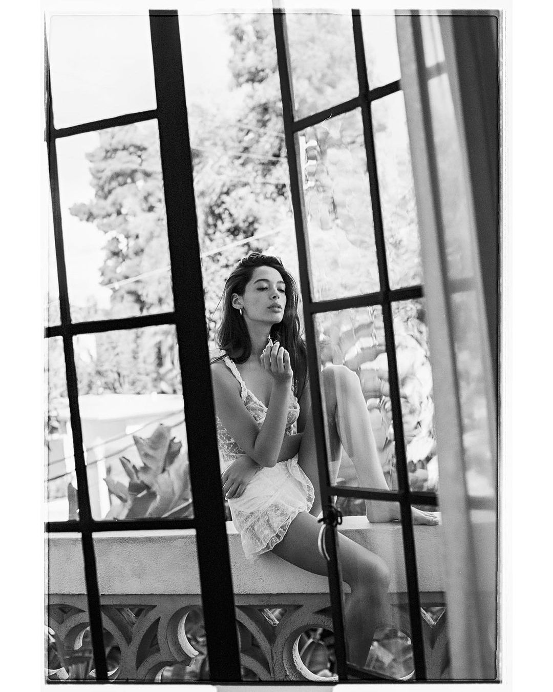 Black and white portrait of a woman in lingerie sitting on a ledge in front of a window with trees visible outside.