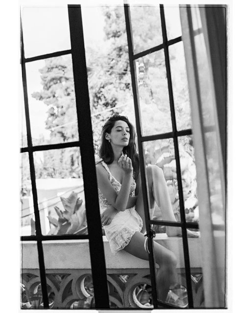 Black and white portrait of a woman in lingerie sitting on a ledge in front of a window with trees visible outside.