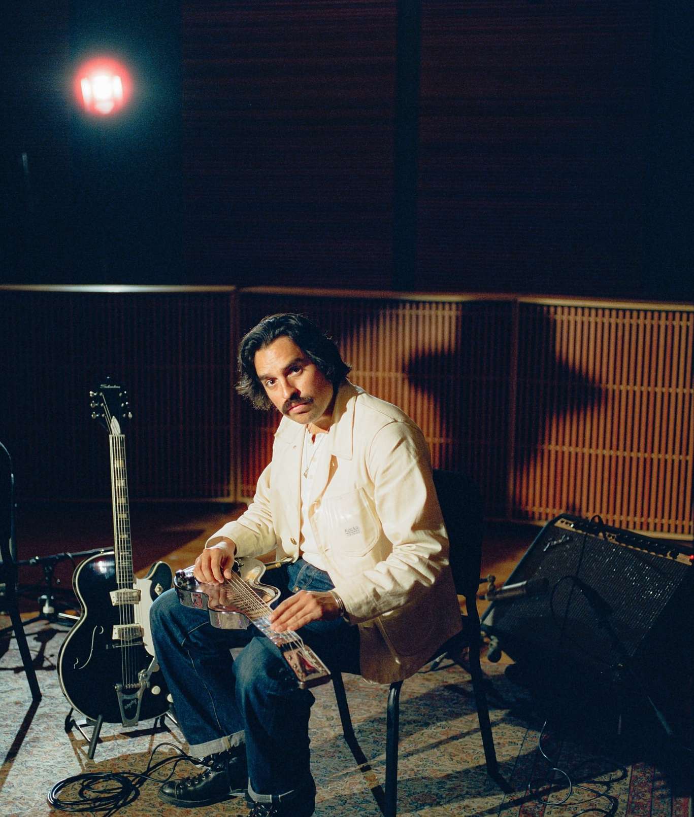 Musician with a mustache sitting in a music studio, holding an electric guitar with a black guitar resting against an amplifier. 