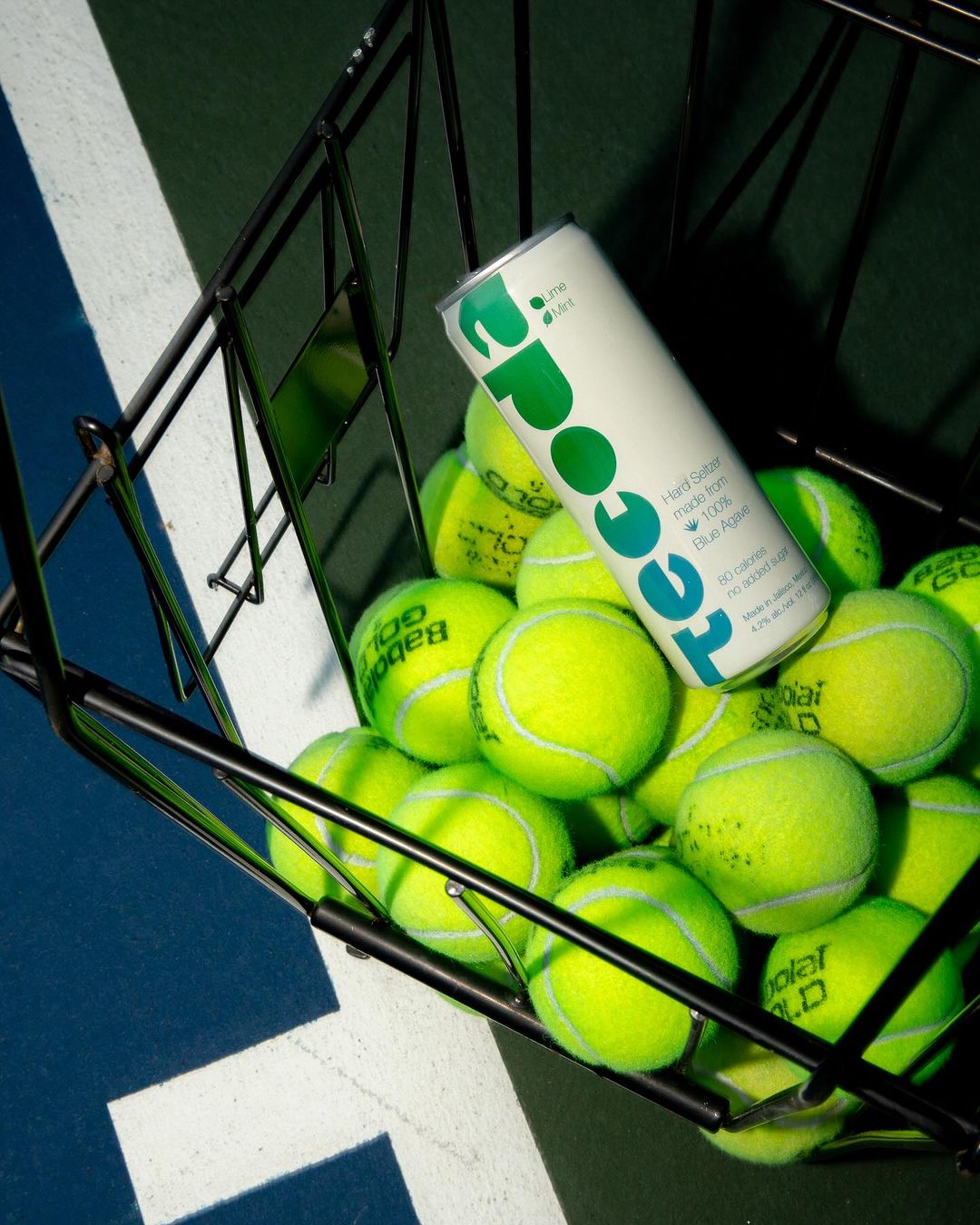 Close-up of a basket overflowing with vibrant yellow tennis balls, featuring a can of hard seltzer against a blue and green tennis court.