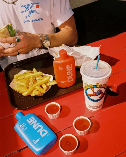 Person eating sandwich at red picnic table with french fries, Dune sunscreen bottles, drink, and ketchup packets in outdoor setting.