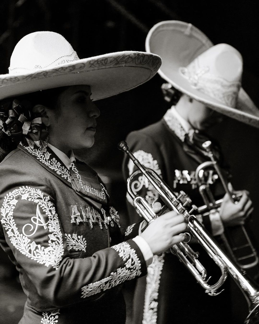 Monochrome portrait of two Mariachi musicians in ornate suits and sombreros playing trumpets in a dimly lit setting.