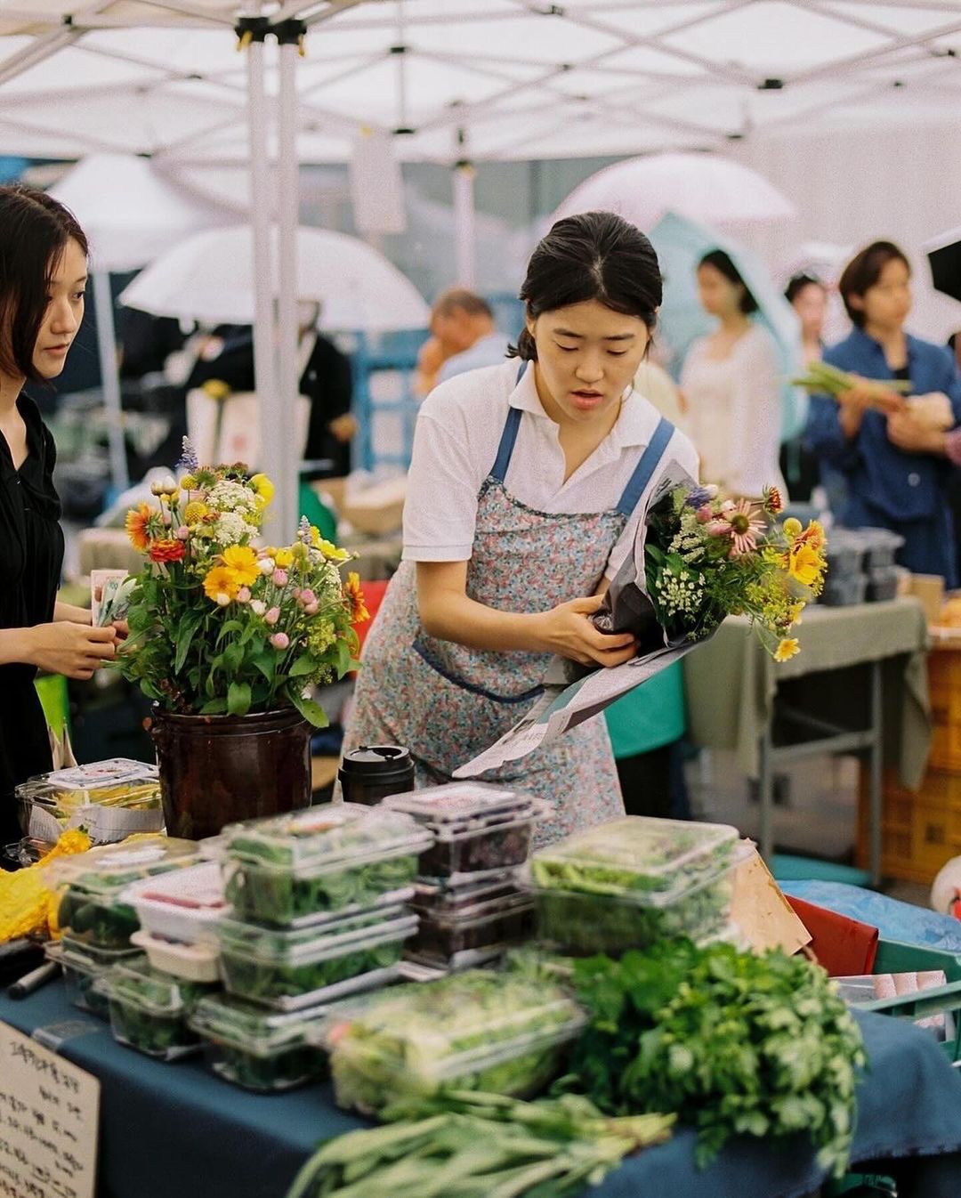 A woman in an apron stands behind a market table selling fresh flowers and produce to a customer on a cloudy day outdoors.
