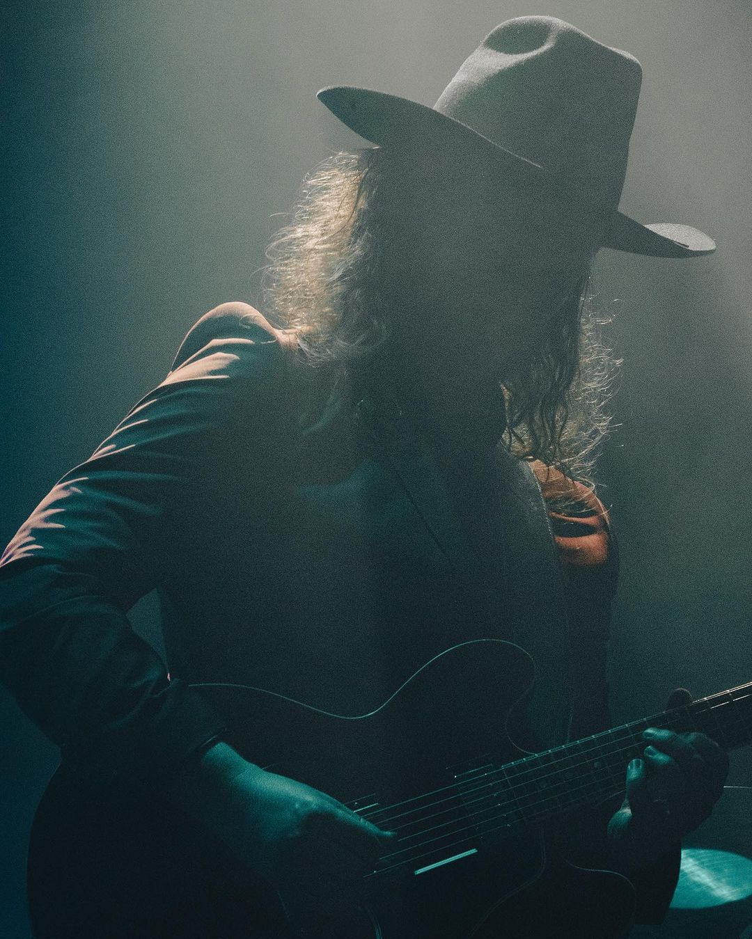 Adult male musician wearing a hat plays an electric guitar in a dimly lit venue with smoky haze for a music or entertainment event.