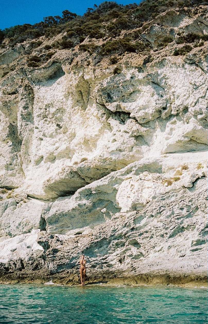 Woman in swimsuit stands near the water's edge at the base of a tall white and tan rock cliff on the shore under a blue sky.
