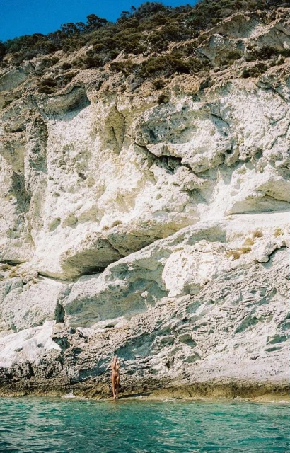 Woman in swimsuit stands near the water's edge at the base of a tall white and tan rock cliff on the shore under a blue sky.