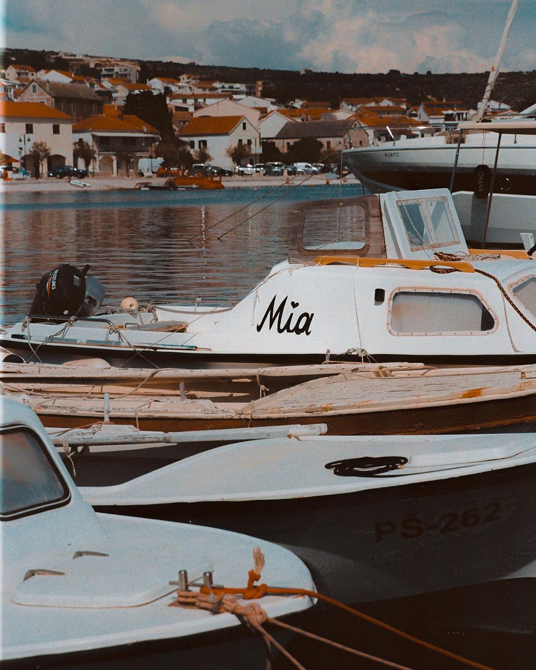 White motorboats docked in a harbor with houses and a hillside visible in the background on a sunny day at a seaside town.