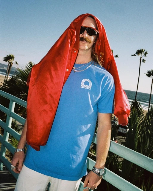 Man with a mustache in a blue tee and sunglasses stands near a beach on a sunny day, wearing a red jacket on his head.