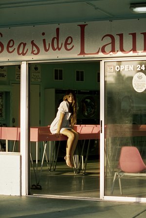 A young woman sits on a pink ledge outside Seaside Laundry, wearing a short dress and sandals in a sunlit, vintage-style photograph.