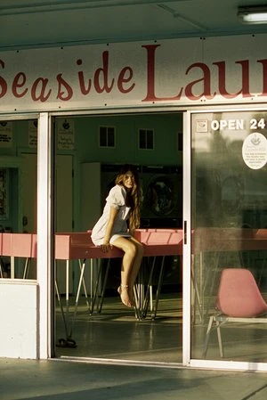 A young woman sits on a pink ledge outside Seaside Laundry, wearing a short dress and sandals in a sunlit, vintage-style photograph.
