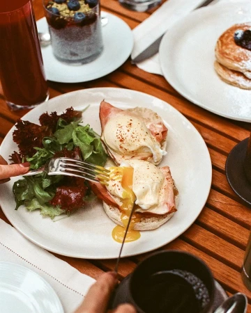 Overhead shot of eggs benedict on a plate with salad, pancakes, and a smoothie on a wood table in a restaurant setting.