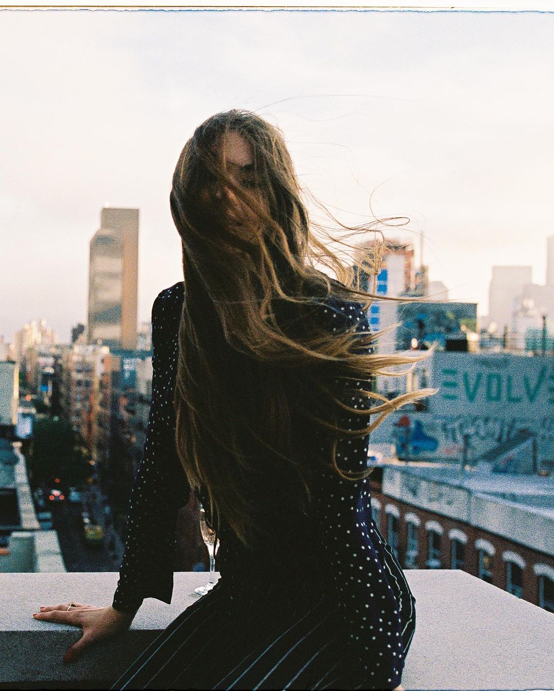 A woman with long hair stands on a rooftop in a polka-dot dress, gazing out over a cityscape under an overcast sky with a building.