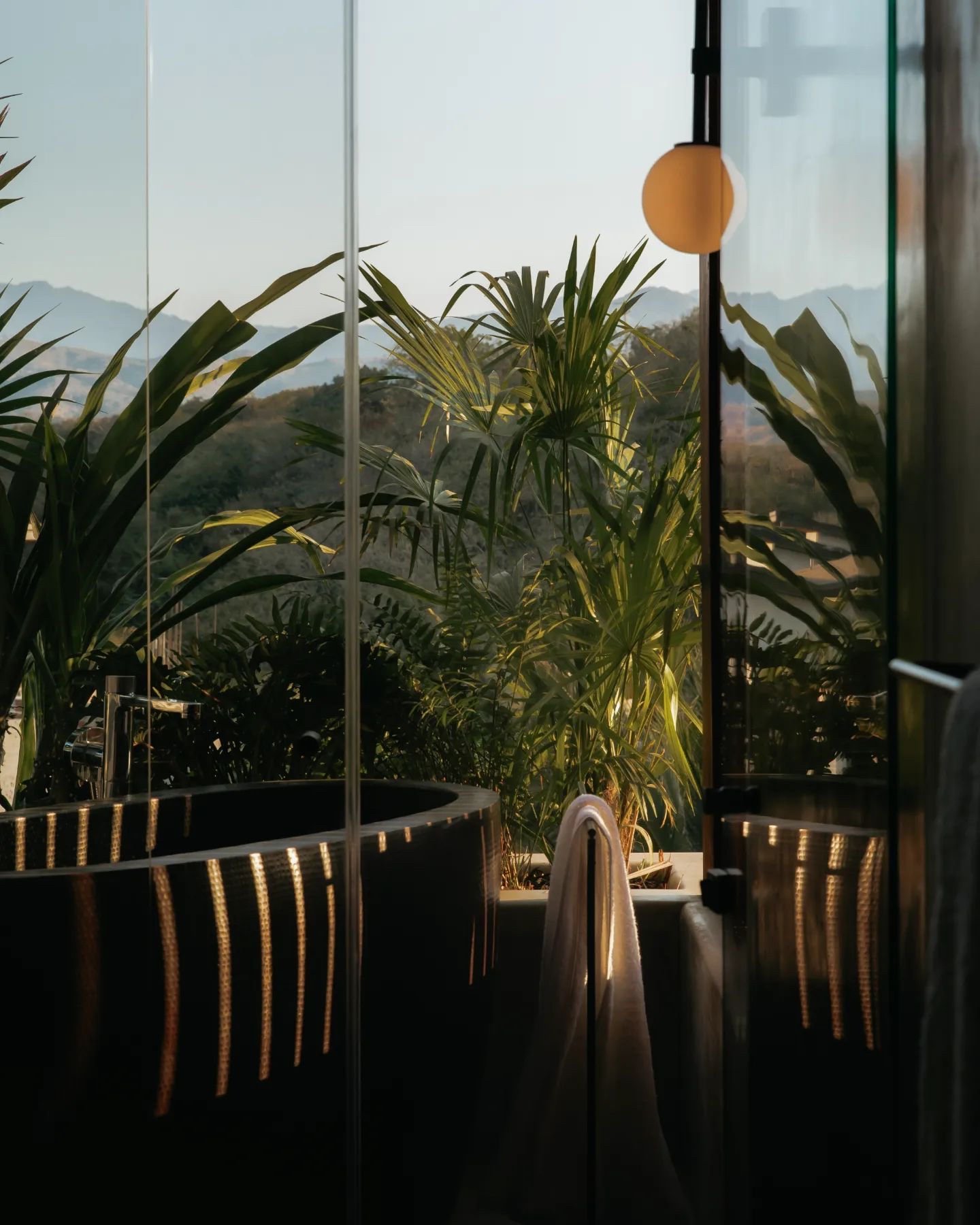 Modern bathroom with a dark tub, a hanging lamp, and large windows overlooking a lush mountain landscape at dusk.