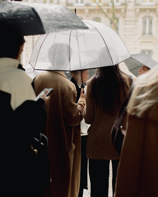 A group of people stand under clear umbrellas on a rainy city street, most wearing coats, in a medium shot emphasizing fashion and urban life.