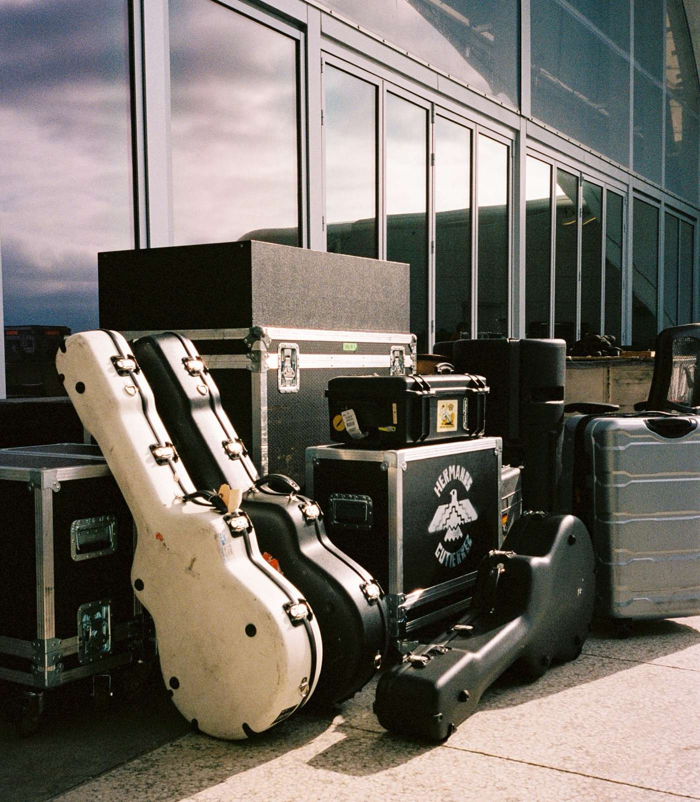 Musical equipment including instrument cases and road cases stacked on a light gray surface in front of large windows.