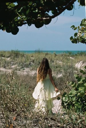 Woman with long brown hair wearing a light yellow tiered dress walking on a sandy beach path framed by coastal foliage