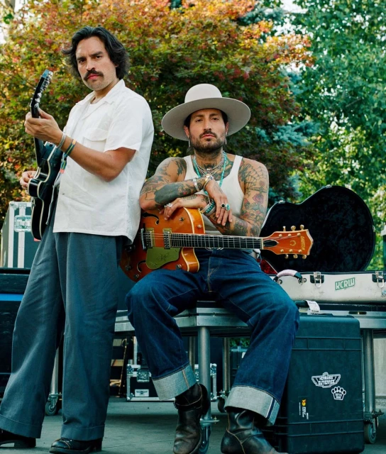Two male musicians outdoors. One plays an electric guitar, wearing a white shirt and grey pants; the other sits with an acoustic guitar.