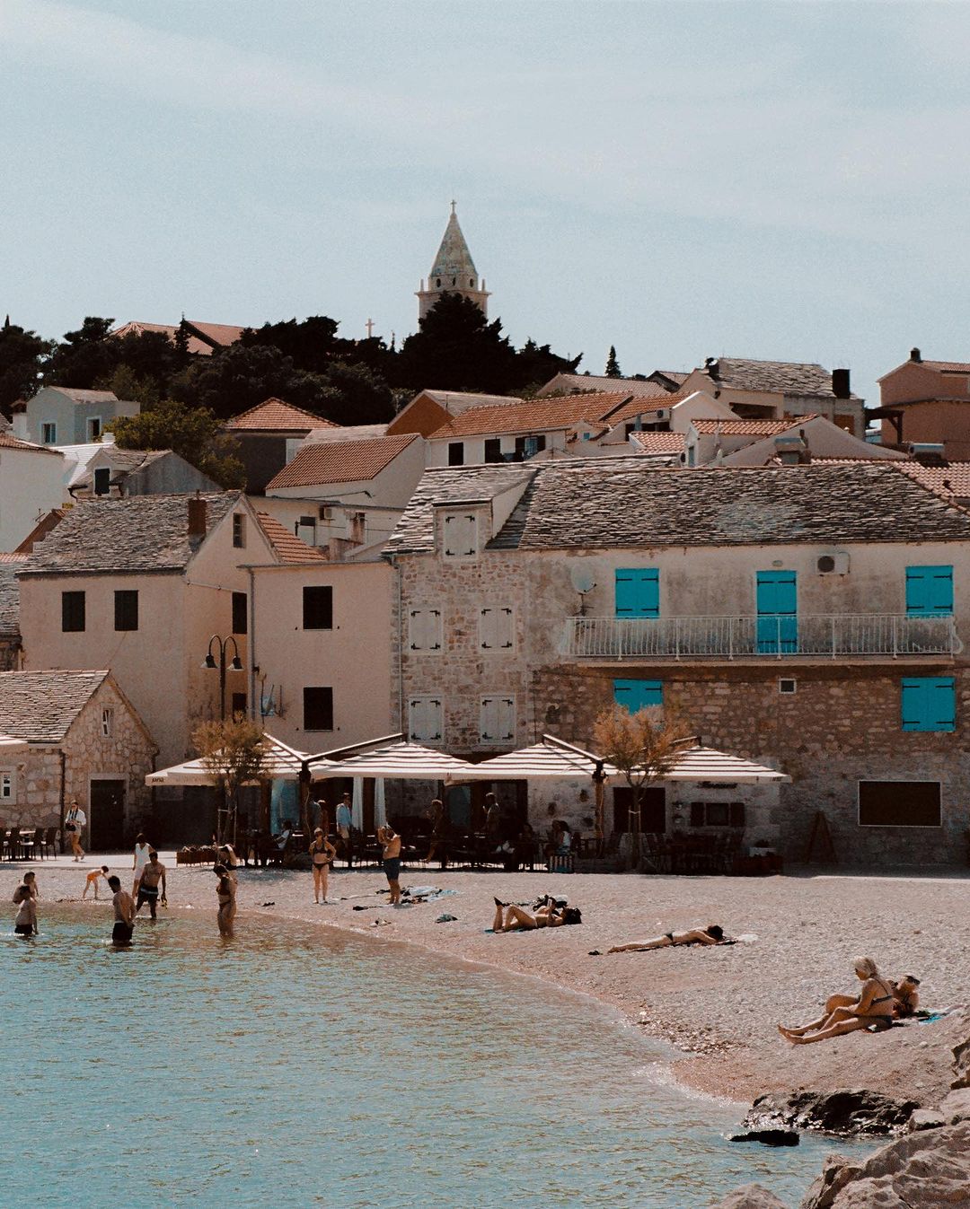 Coastal town with stone buildings, beachgoers enjoying the pebble beach, and clear turquoise water on a sunny day near the old village.