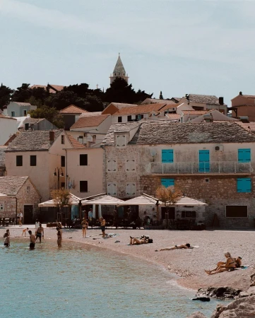 Coastal town with stone buildings, beachgoers enjoying the pebble beach, and clear turquoise water on a sunny day near the old village.