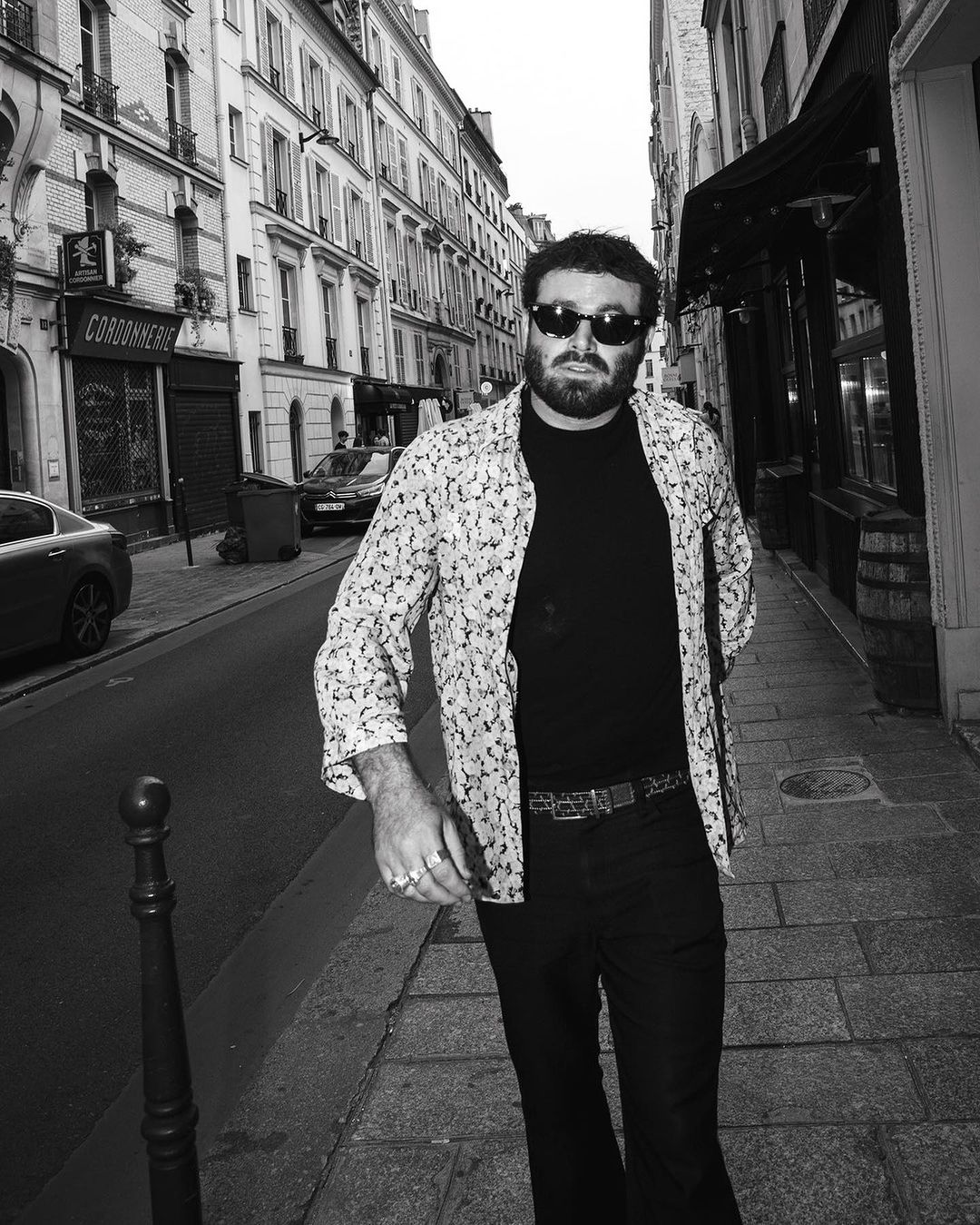 Black and white street-style portrait of a man with sunglasses in an open patterned shirt on a Parisian street.